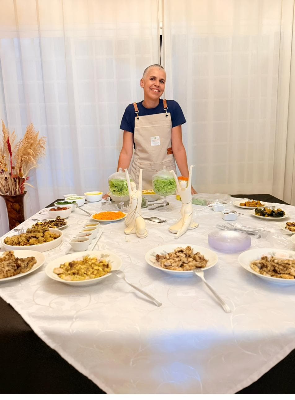 Leila Campelos sorrindo atrás de uma mesa posta com travessas das suas receitas funcionais